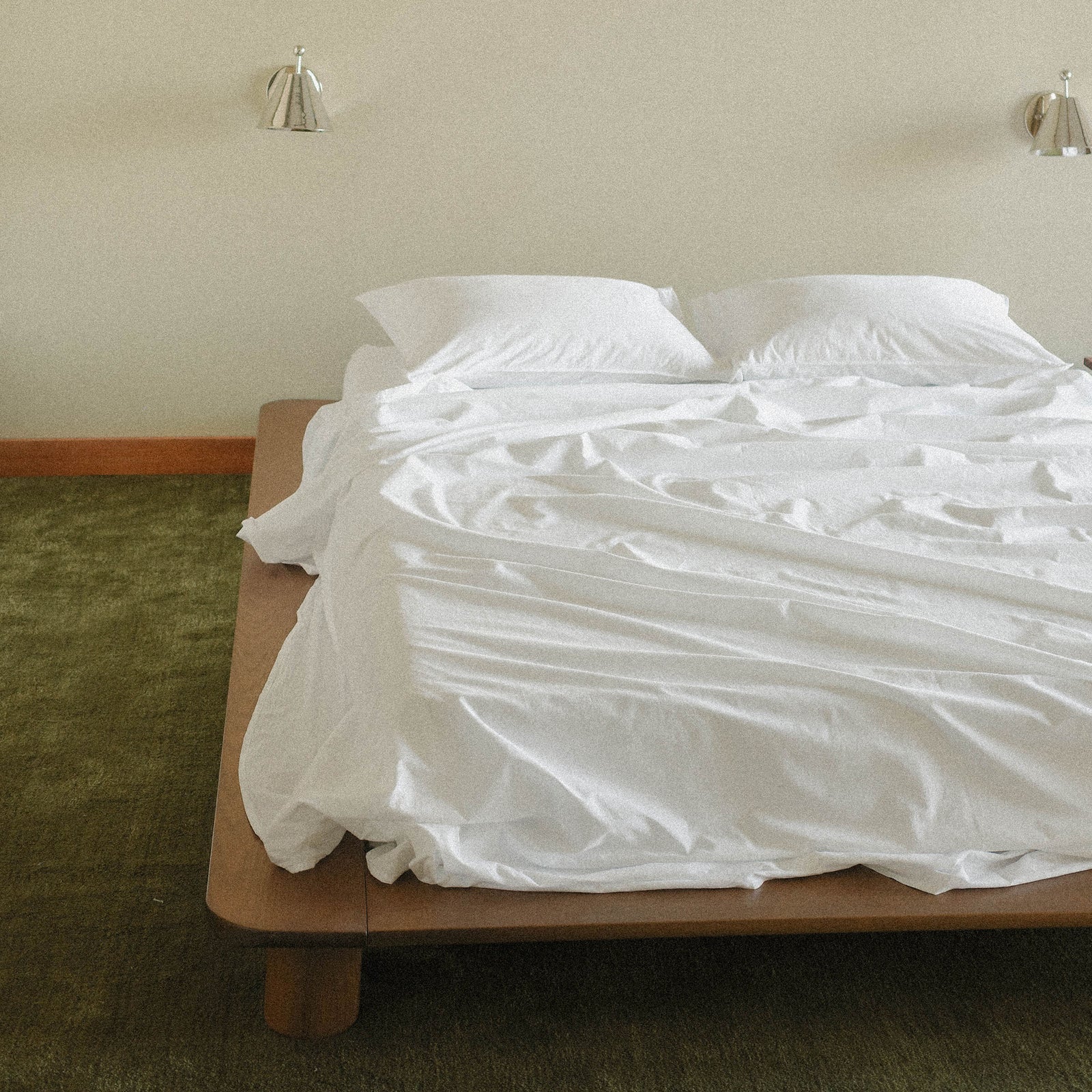 Bed with white bedding in a room with beige walls, two silver sconces and green carpet.