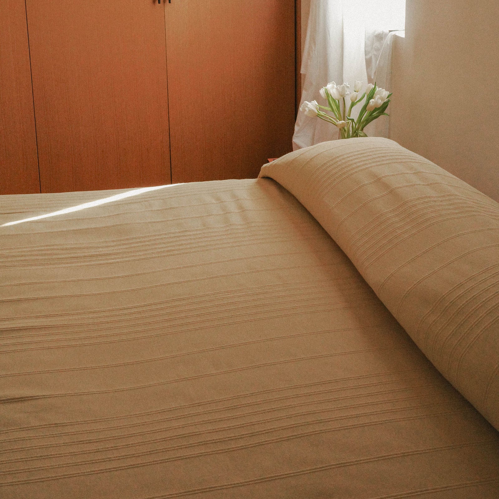 Beige / yellow bedspread on a bed with a wooden wardrobe and a vase of white tulips in the background