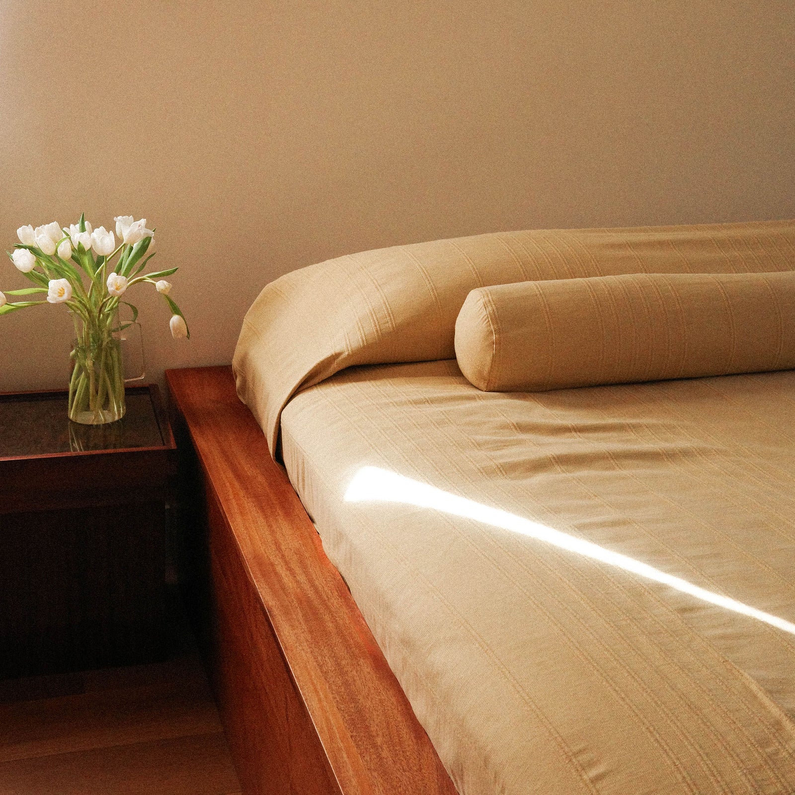 A bed with beige / yellow sheets beside a side table with flowers against a beige wall. There is a bolster on the bed. 