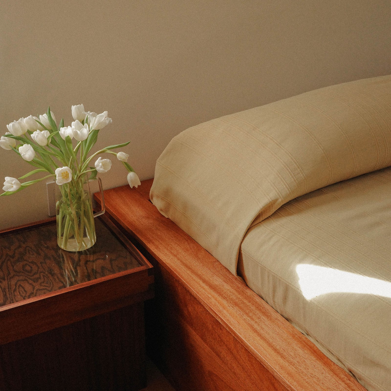 Bed with beige / yellow bedding and a vase of white tulips on a wooden nightstand.