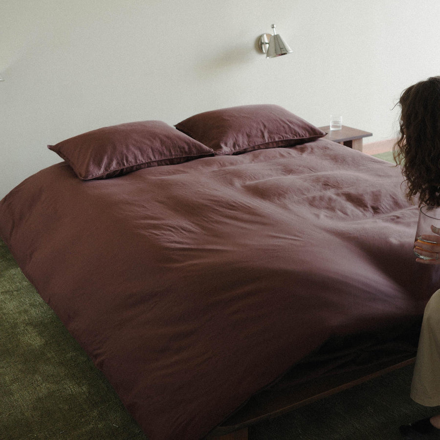 A neatly made bed showing a duvet cover in purple / brown with two pillows in purple / brown. A person is pulling the bed taught. There is a green carpet