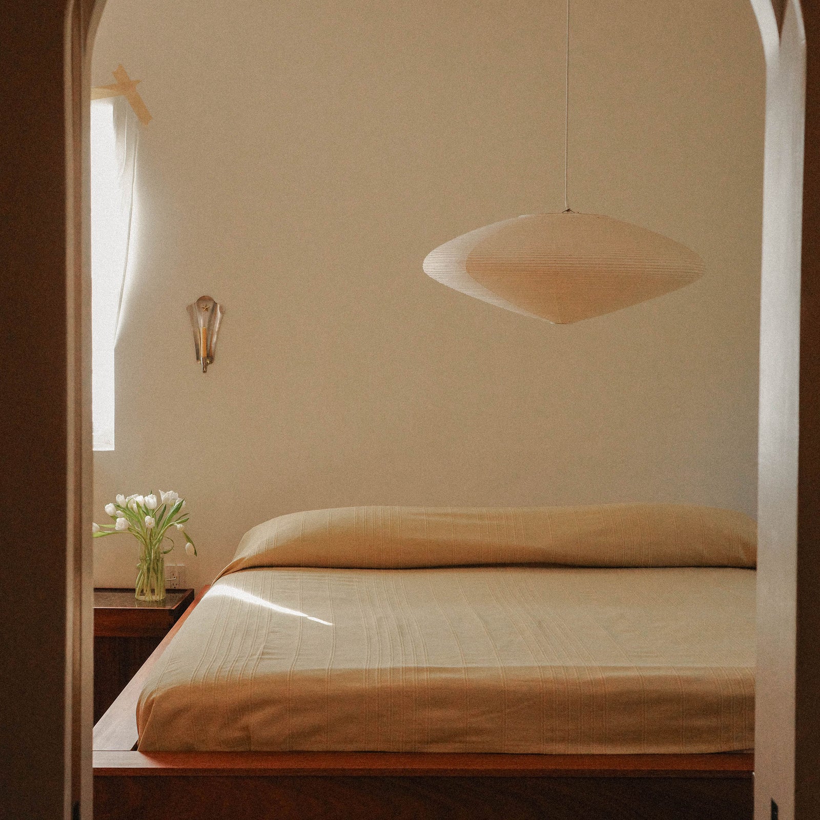 Bedroom with beige walls, a bed with beige/ yellow sheets, and a pendant light.