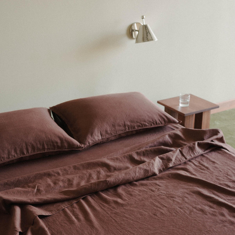 A bed dressed in brown / purple sheets, with two pillows. The bed is beside a wooded nightstand with a glass of water beneath a single chrome sconce