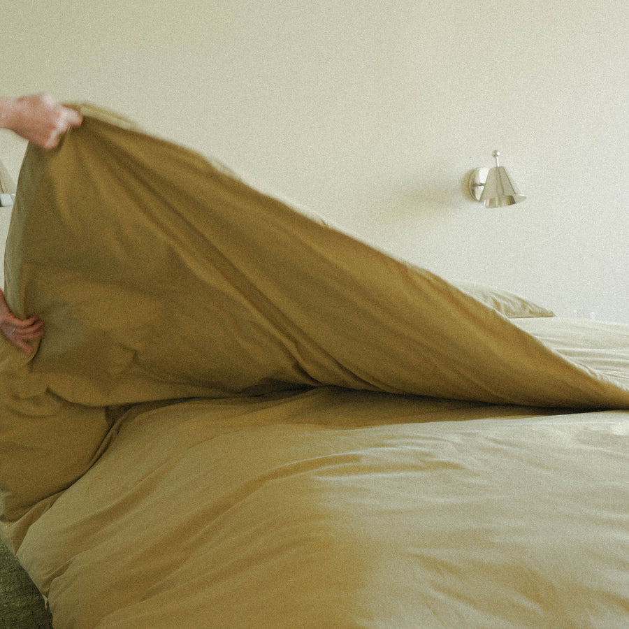 Person making a bed with green duvet cover in a room with a silver sconce on the wall.
