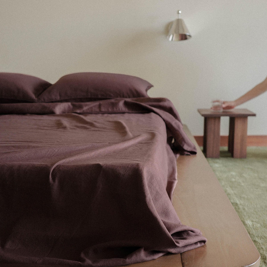 A wood bed with brown / purple sheets in a bedroom setting - there is a hand placing a glass of water and a green rug