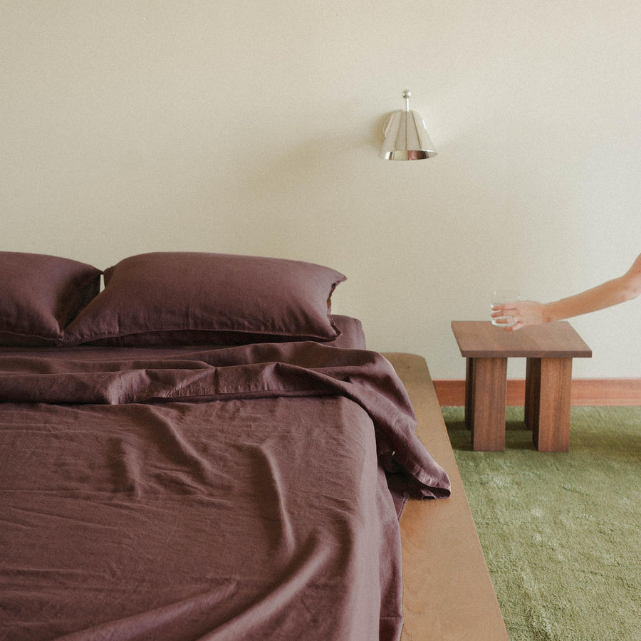 A wood bed with purple / brown sheets and two pillows. There is a chrome sconce hanging over a wooden nightstand with a hand placing a glass of water down on the nightstand. 