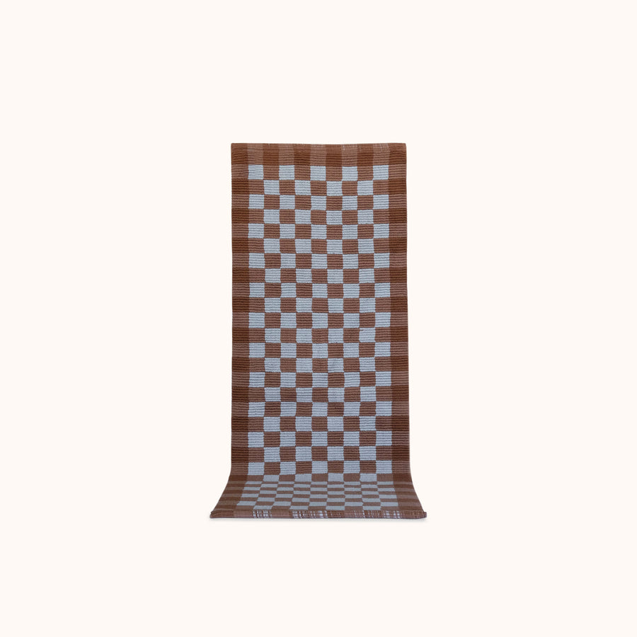 A long rectangular woven mat with a brown and light blue checkerboard pattern, displayed flat against a solid off-white background.