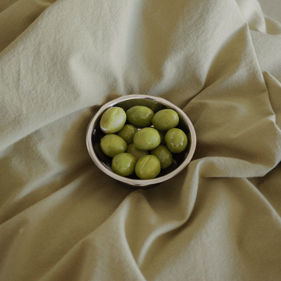 A small metal bowl filled with glossy green olives, placed on draped light green fabric.