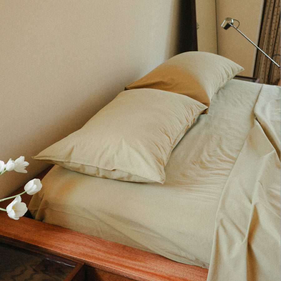 Light green cotton bed sheets and matching pillowcases on a bed with a wooden headboard, styled with white tulips and a bedside lamp.