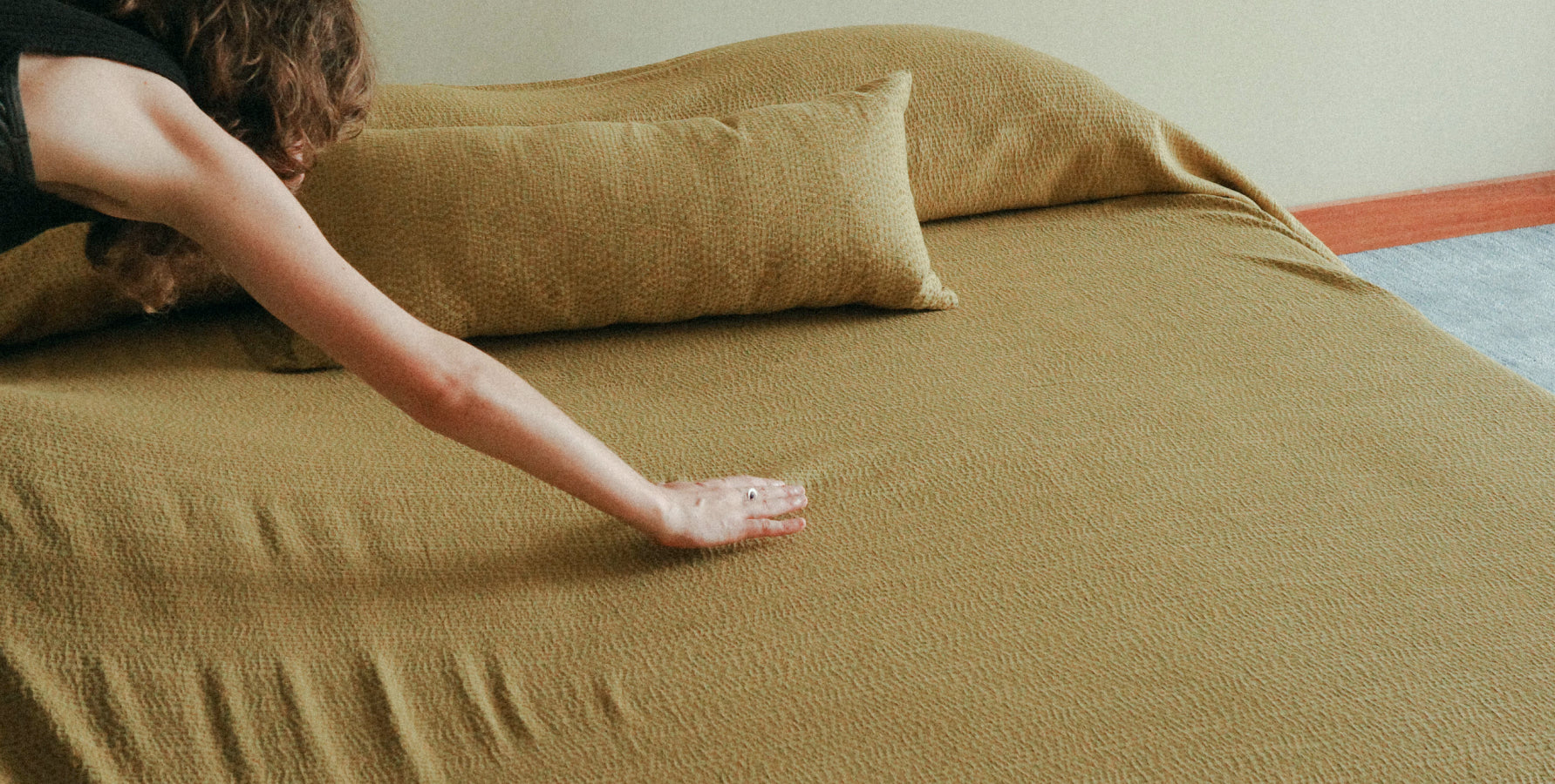 A person's arm and hand stretching a textured olive green bedspread over a bed with matching pillows in a bedroom setting.