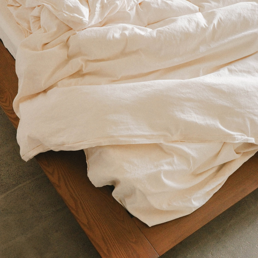 A close-up of a light cream or off-white bedding fabric, likely a duvet cover or comforter, draped over a wooden bed frame on a concrete floor.