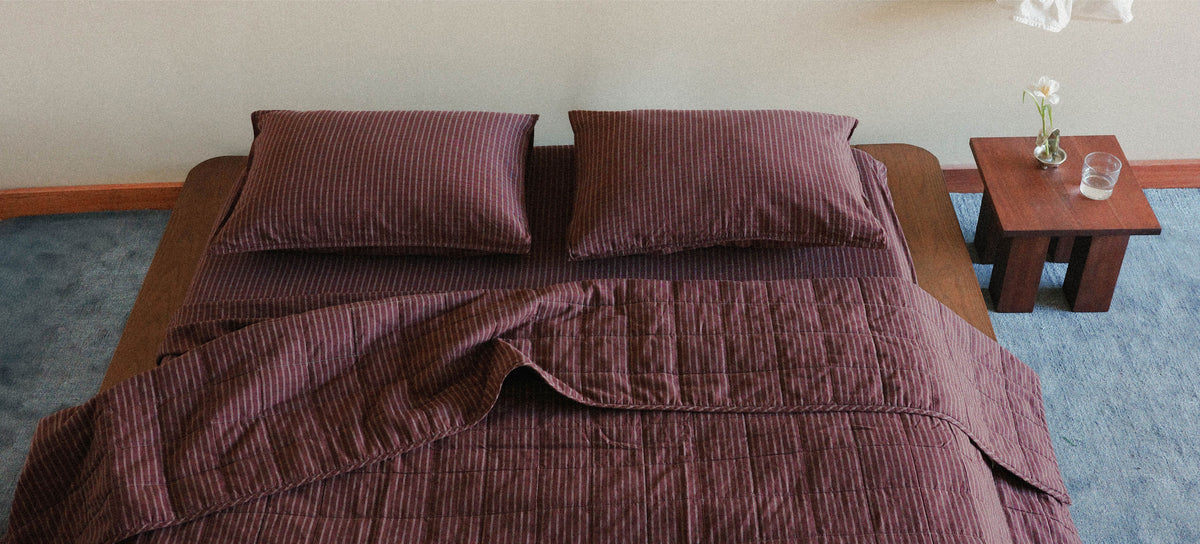 A bed dressed in a dark maroon check bedding set with two matching pillowcases, shown in a styled bedroom scene with a small wooden side table and blue carpet.