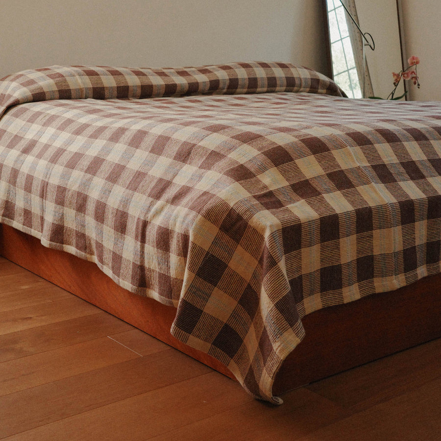 A bed dressed in a brown and beige plaid check blanket, shown in a bedroom with a wooden floor and a window in the background.
