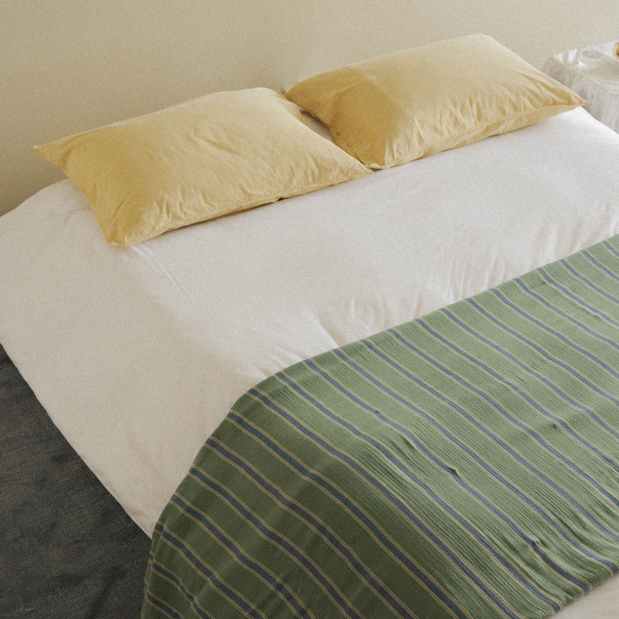 A neatly made bed with white bedding, two pale yellow pillows, and a green striped throw blanket draped across the corner.
