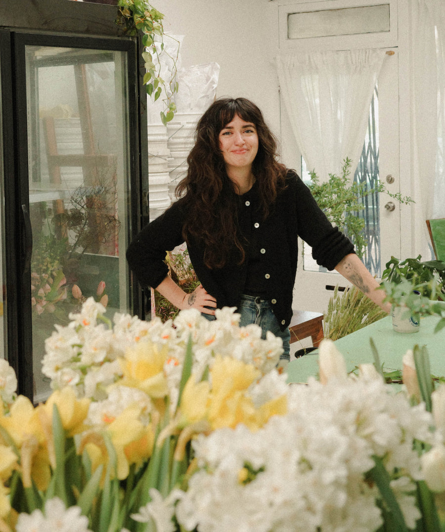 A woman stands in a bright flower shop next to a large arrangements of white and yellow flowers fill the foreground, with plants and glass display cases visible in the background.