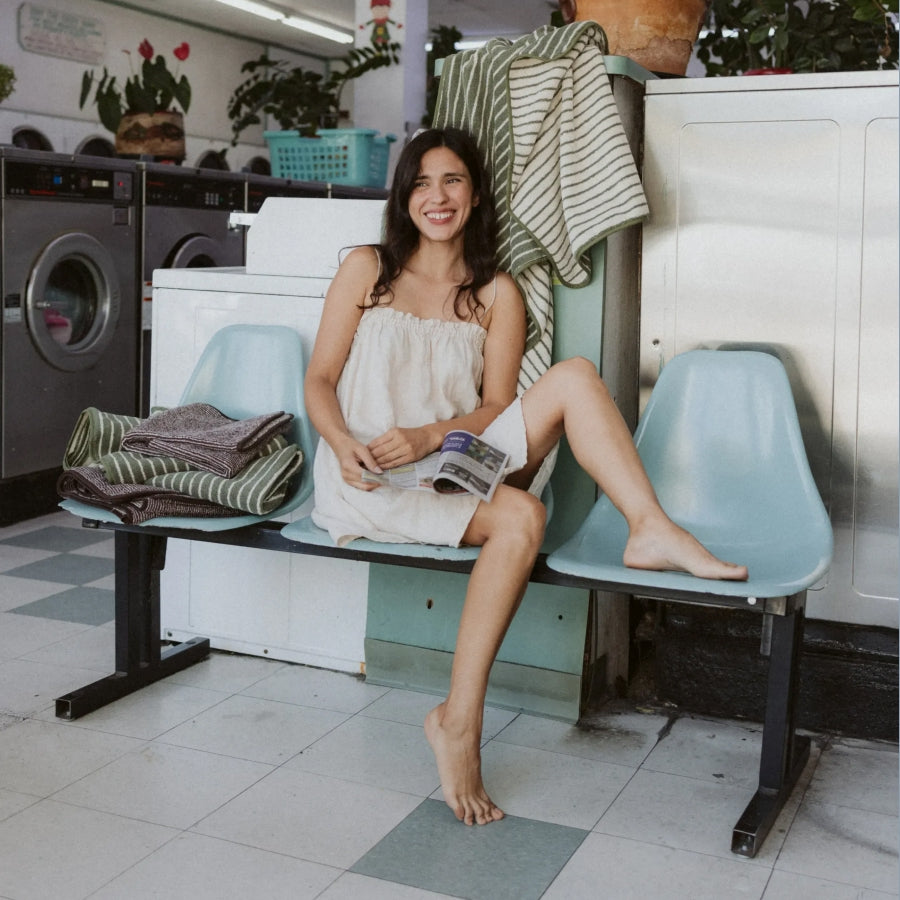 A woman smiling while sitting on a light blue bench in a laundry room, wearing a white strapless dress and holding a magazine. Laundry items and washing machines are visible in the background.