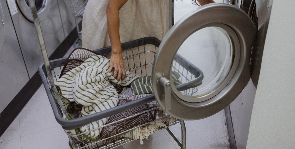 A person in a beige dress pushes a metal laundry cart filled with striped towels and clothes toward a washing machine in a laundry room.