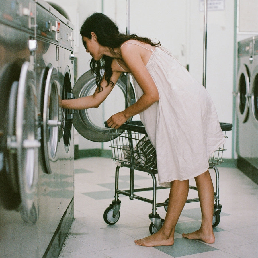 A woman in a white nightgown leans over a metal laundry cart next to a large washing machine.