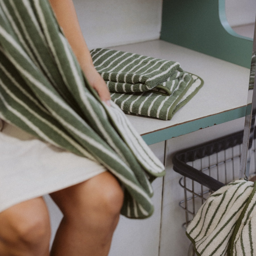A person holding a green and white striped bath towel next to a matching folded towel on a light-colored table.
