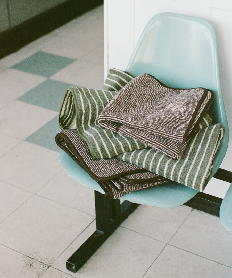 A stack of folded textured knit blankets and towels in muted green, brown, and cream stripes, resting on a light blue plastic chair in a tiled indoor space.
