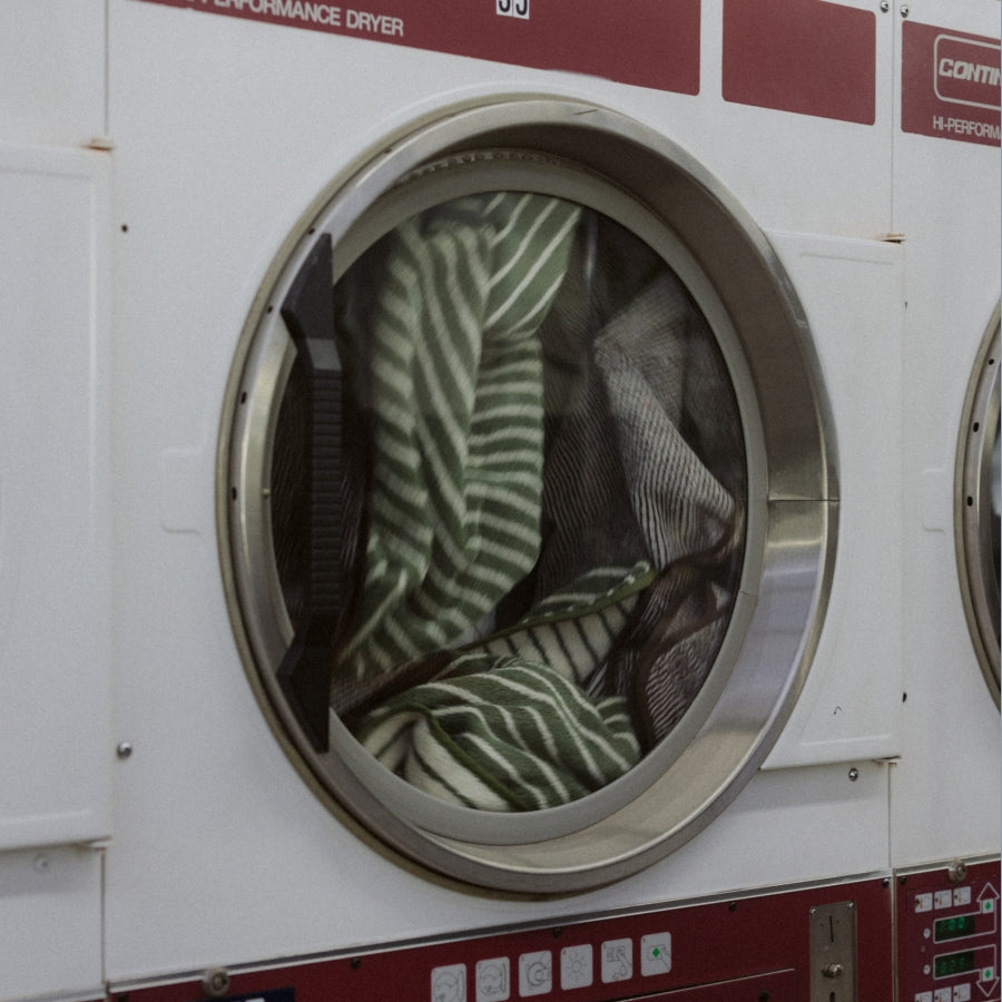 A close-up of a white and maroon industrial laundry machine door with a circular window, showing green striped towels inside.