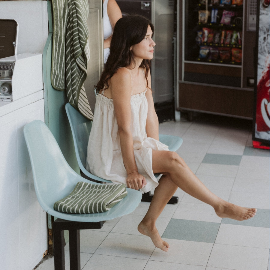 A woman sits on a mint green molded chair in a Lavandería, wearing a white sleeveless dress. A striped green and white towel rests on the chair next to her.
