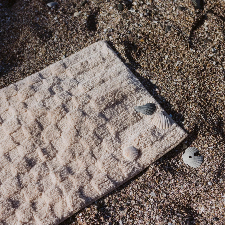 A pink bath runner with alternating shag heights on a sandy beach with 3 seashells on top.
