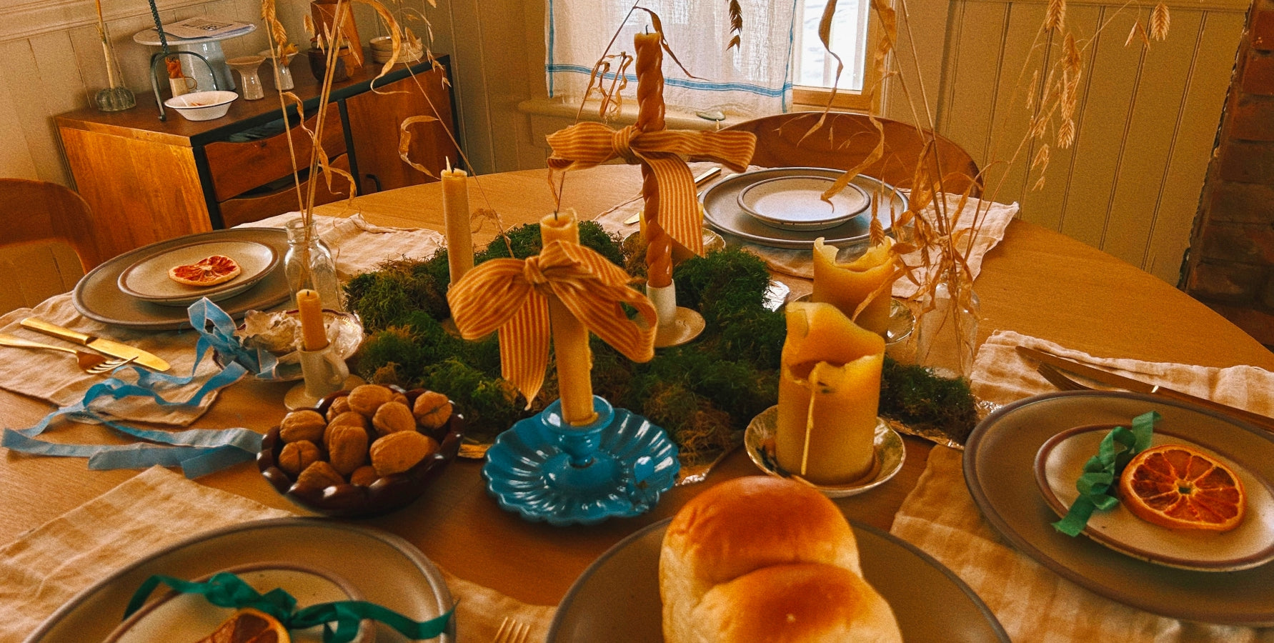 A warmly lit dining table set for a meal, featuring round plates, bread rolls, olives, decorative blue dishes, dried grasses, and a central green garland centerpiece.