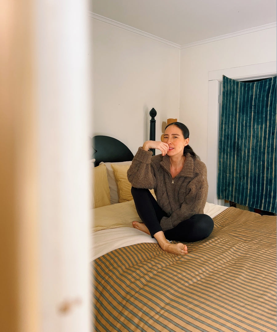 A woman sits cross-legged on a bed in a bedroom. The bed has a striped tan and brown comforter, and a blue-green striped curtain hangs on the window behind her.