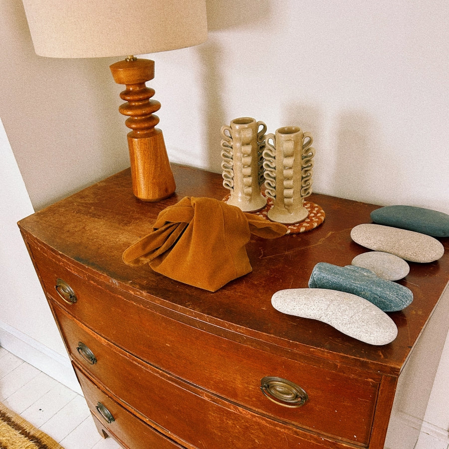 A vintage wooden chest of drawers with brass handles, styled with a wooden table lamp, two cream ribbed ceramic candle holders, a folded mustard-brown fabric bag, and several smooth decorative stones.