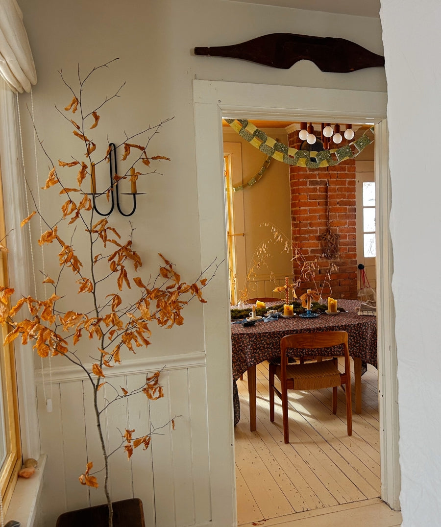 A rustic autumn-themed interior scene featuring a tall decorative tree branch arrangement with dried orange-brown leaves on the left, set in a doorway that leads to a dining room with a table, chairs, and a brick wall.