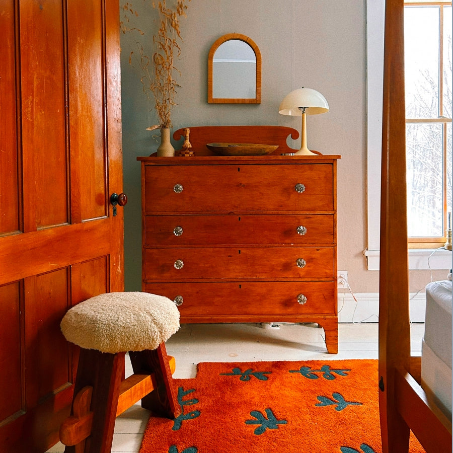 A mid-century modern wooden chest of drawers in a warm reddish-brown finish, styled in a bedroom setting with a small stool, orange rug, and window.