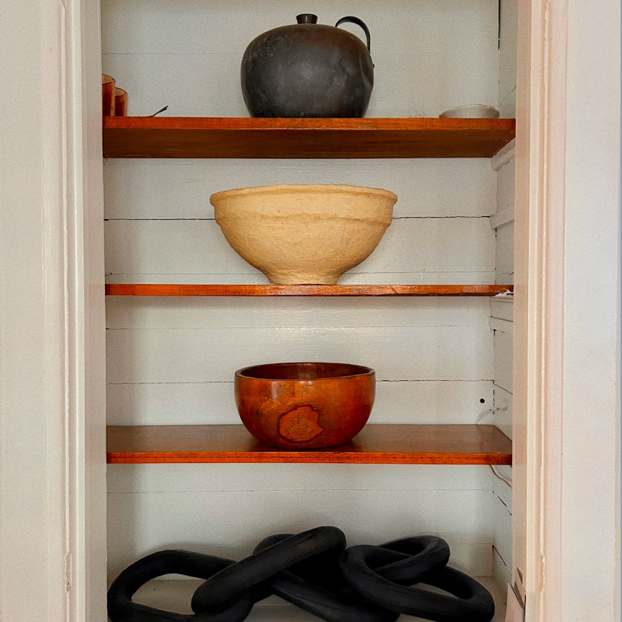 Three rustic wooden bowls displayed on shelves inside a white pantry or storage closet, with a dark ceramic pot on the top shelf and black coiled decor on the bottom shelf.