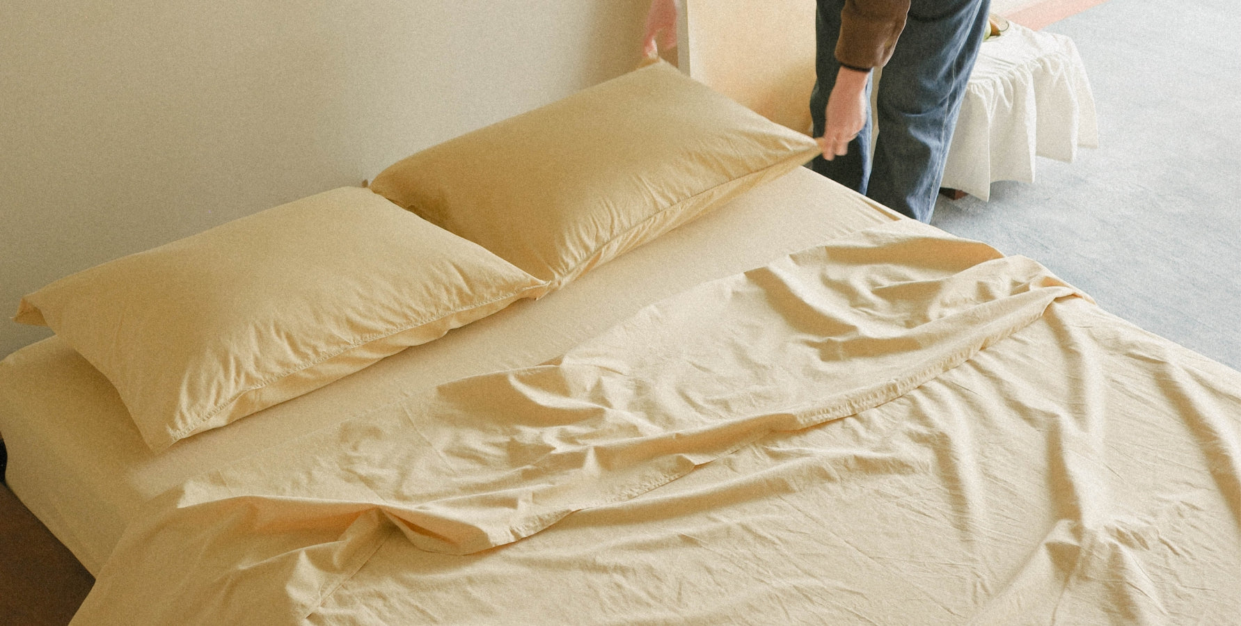 A bed dressed in light yellow bedding, including two pillows and a rumpled sheet, with a person's hands and legs visible adjusting the top corner of the duvet in a softly lit room.