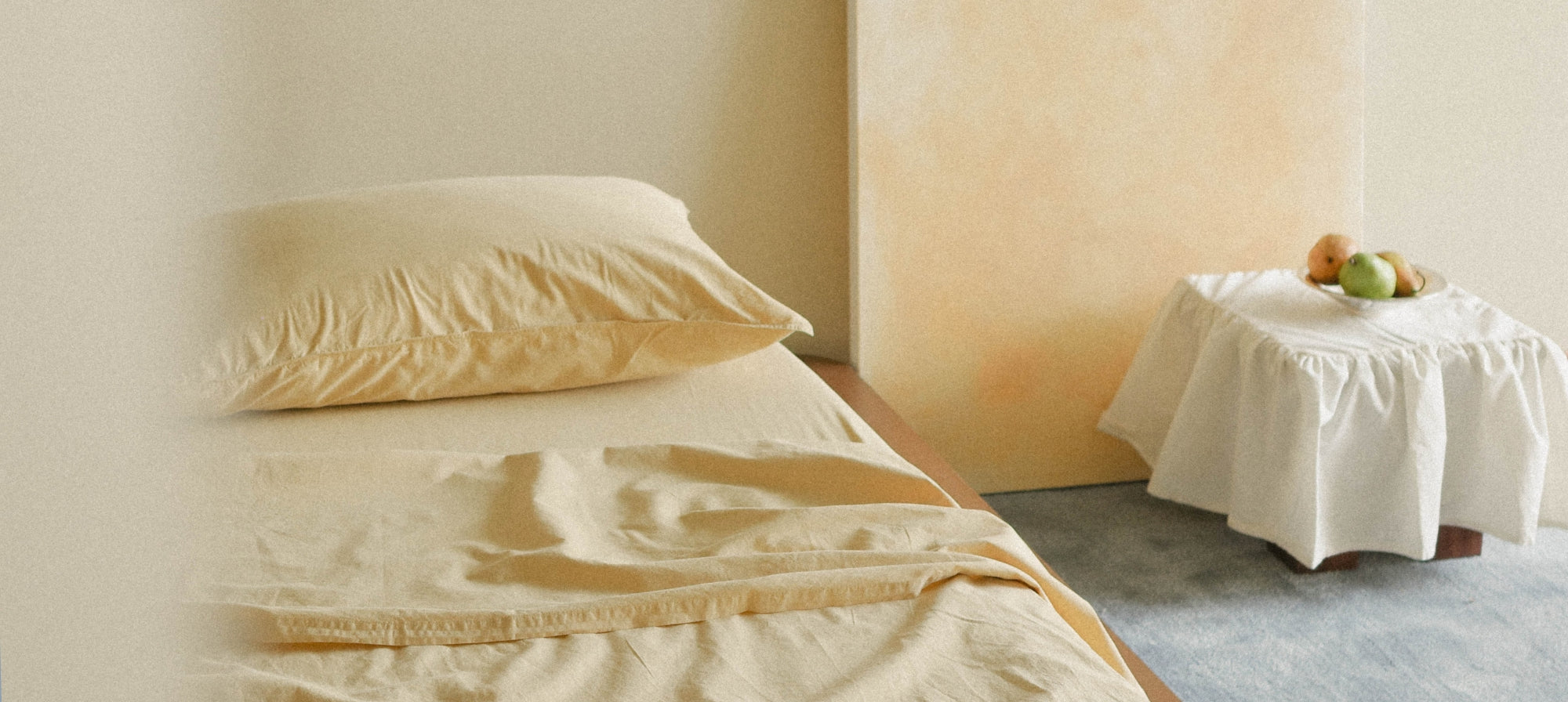 A close-up of a bed dressed in light yellow bedding, with a matching pillow and rumpled sheet, in a warm-toned bedroom setting. A small draped stool with fruit sits in the background.