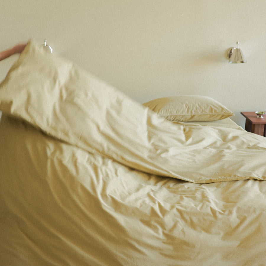 A person's hand is visible at the left edge as they lift a pale olive duvet cover over a bed in a minimalist bedroom setting.