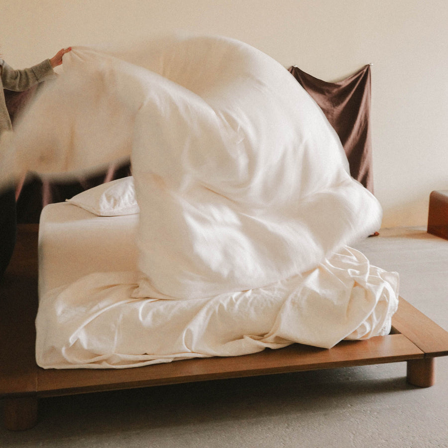 A person's hand and arm are visible as they lift and spread a large, soft white duvet cover over a bed on a wooden platform bed frame in a minimalist bedroom setting.