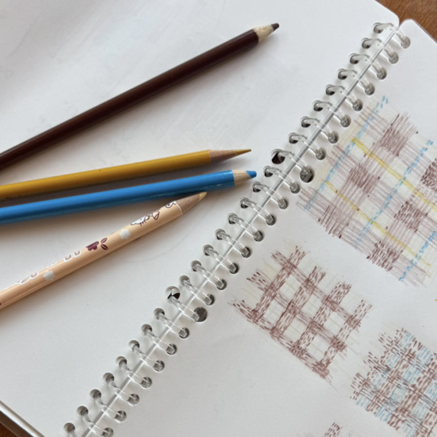 An open sketchbook showing brown check patterns next to an assortment of colored pencils 