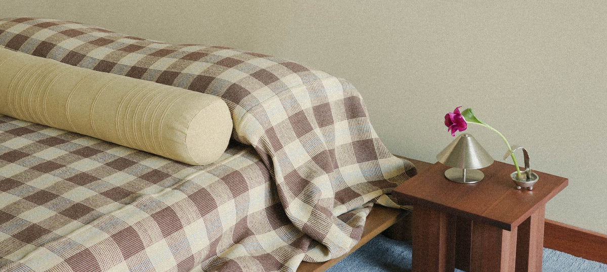 Brown and beige check blanket draped over a bed with a long yellow bolster pillow next to a low wooden side table with a small lamp and pink flower.