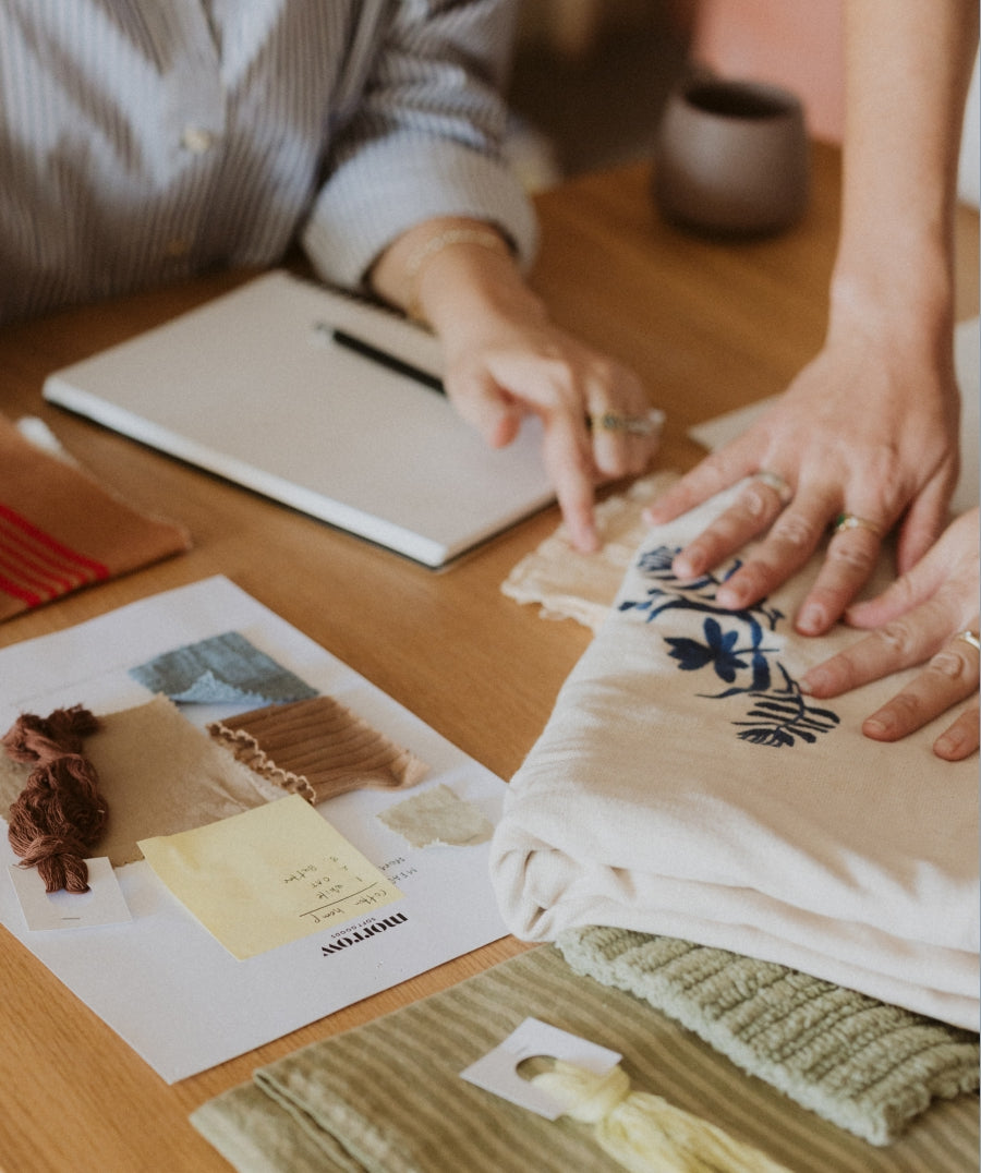 A close-up lifestyle shot of a person's hands folding a cream-colored fabric, set on a wooden desk alongside fabric swatches and a notebook.