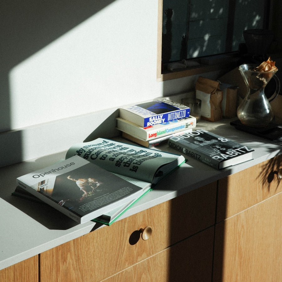 A styled interior scene with a light wood cabinet and countertop in strong sunlight. Several books are stacked and spread out on the counter.