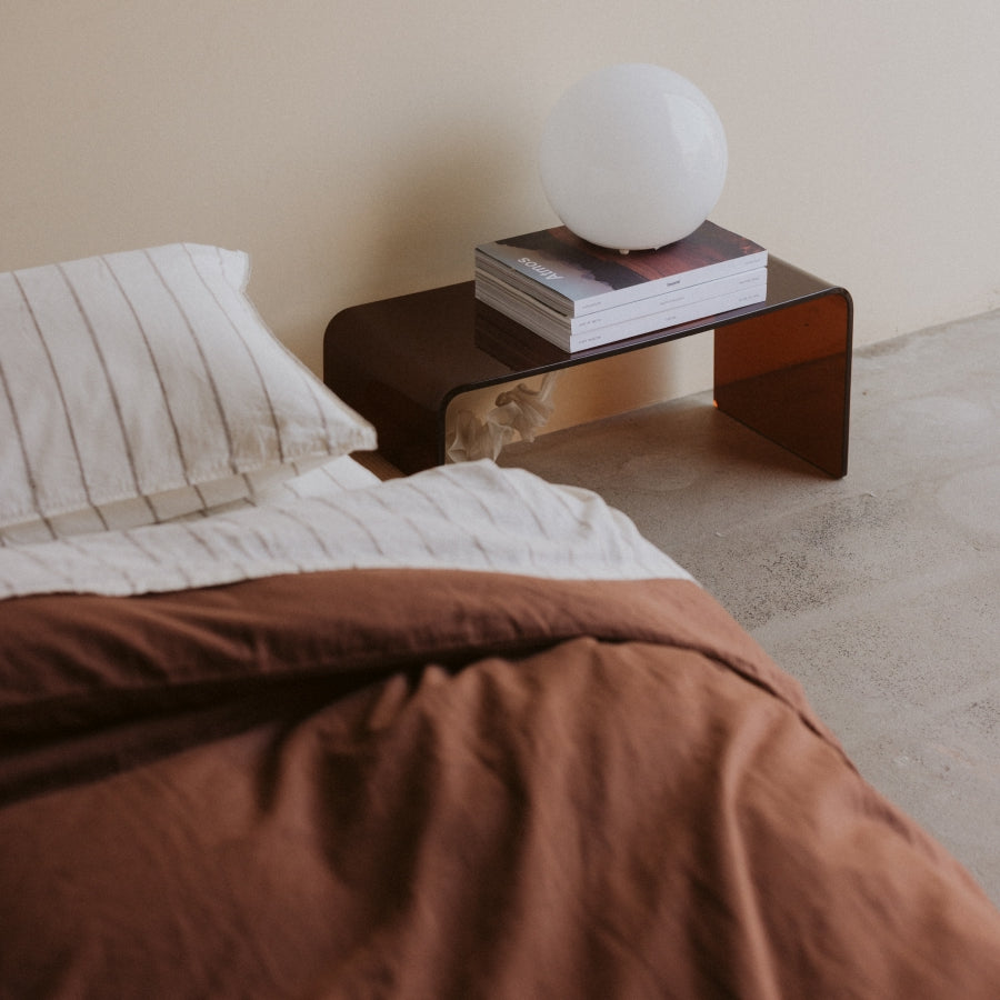 A modern translucent dark brown acrylic side table in a bedroom setting, styled with a white globe lamp, books, and a striped pillow on a bed with rust-colored bedding.