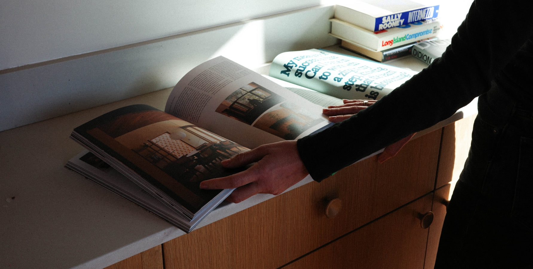 A person in a black long-sleeve top holds an open magazine on a light-colored wooden cabinet, with other books stacked on top in a sunlit room.