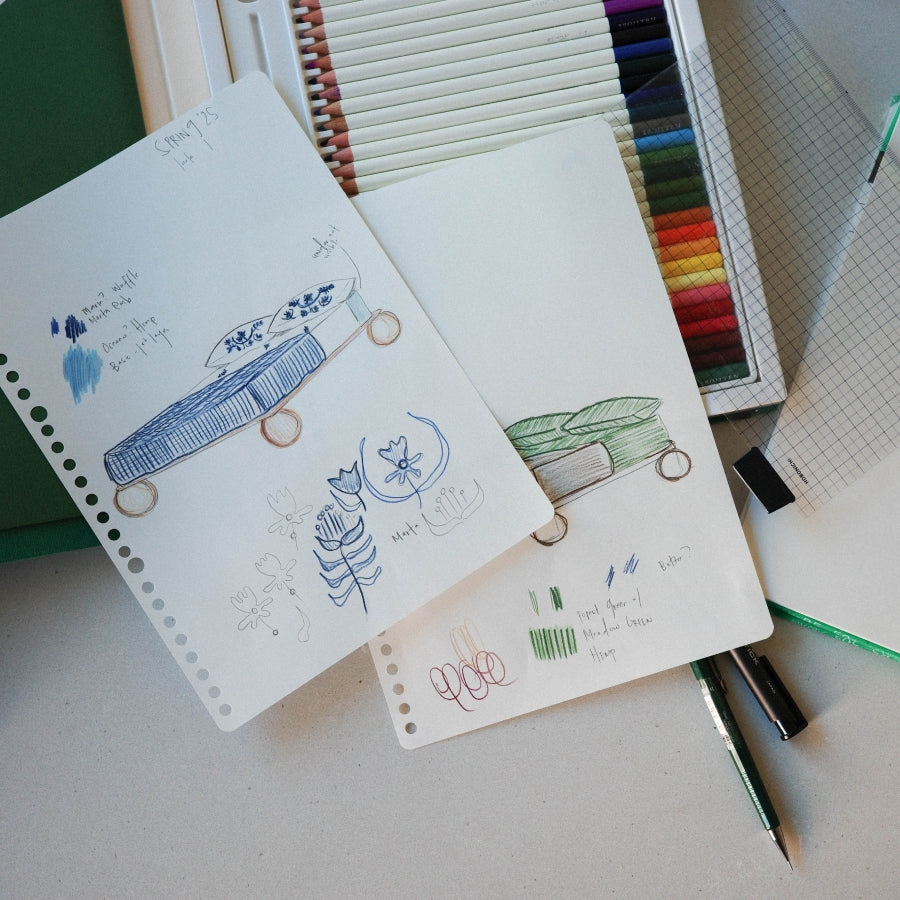 Three spiral-bound sketch notebooks with hand-drawn illustrations of a beds, arranged on a desk with colored pencils, a grid sheet, and pens.