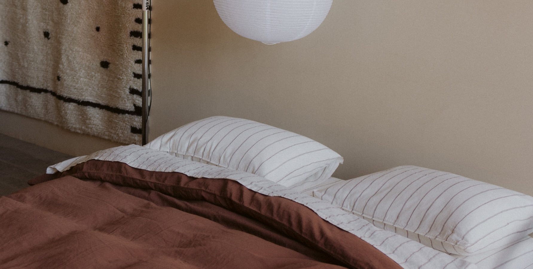 A cozy bed setup with two white striped pillows and a brown duvet cover, set against a beige wall with a hanging textile and a white hanging light fixture.