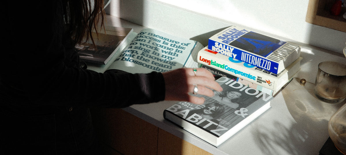 A person in a black top reaches toward a stack of books on a light-colored countertop in a real indoor setting with strong sunlight and shadows.