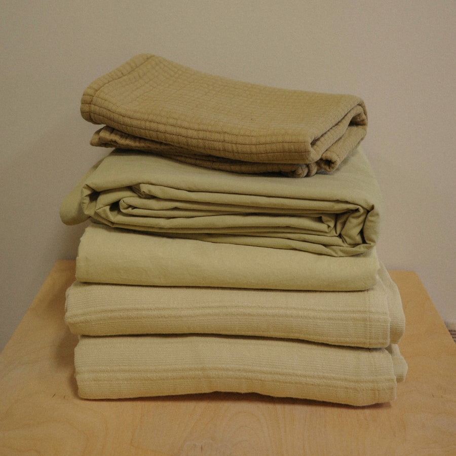 A stack of five folded textiles in varying shades of beige and light olive green, arranged on a light wood surface against a plain beige wall.