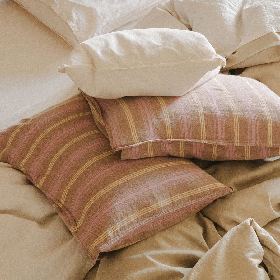 Three pillowcases arranged on a bed: two have a muted pink and purple stripe pattern with thin yellow lines, and one is solid cream. The pillows are styled on beige bedding.