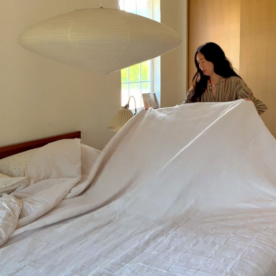 A woman spreads a large white quilt across a bed in a softly lit bedroom. A hanging pendant lamp, window, and wooden wall are visible in the room.