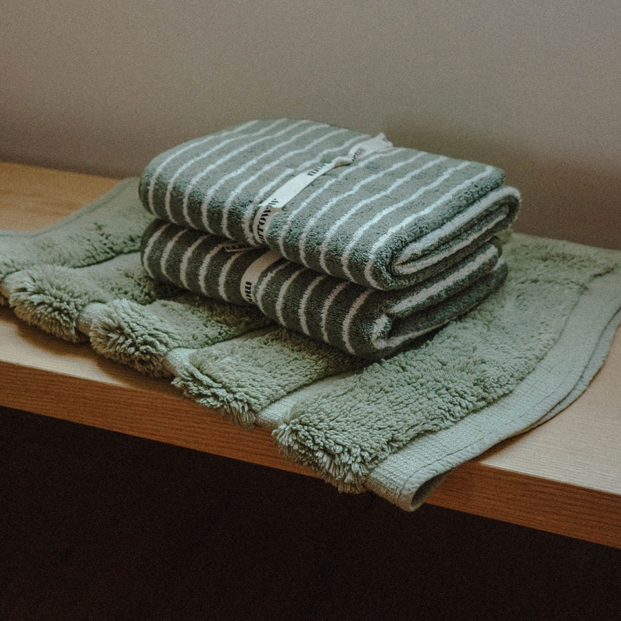 A stack of folded bath towels in sage green and white stripes, placed on top of a folded matching bath mat on a wooden shelf against a neutral wall.